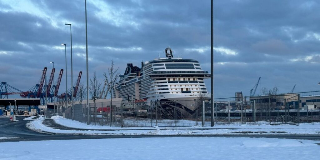 A winter scene showing a snow-covered ground with a large MSC cruise ship docked at a busy port. Industrial cranes and a partly cloudy sky create a contrasting backdrop to the modern luxury liner in an otherwise industrial environment.