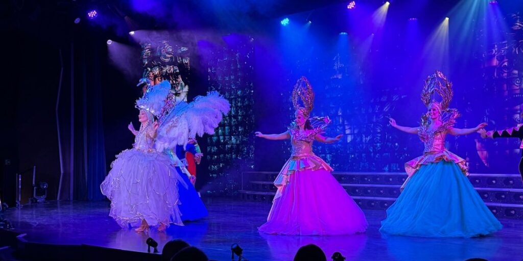Performers in elaborate costumes take the stage during a vibrant show on a cruise ship. Three dancers, adorned with large feathered headpieces and voluminous, pastel gowns, are illuminated by dramatic stage lighting, creating a lively and enchanting spectacle for the audience.