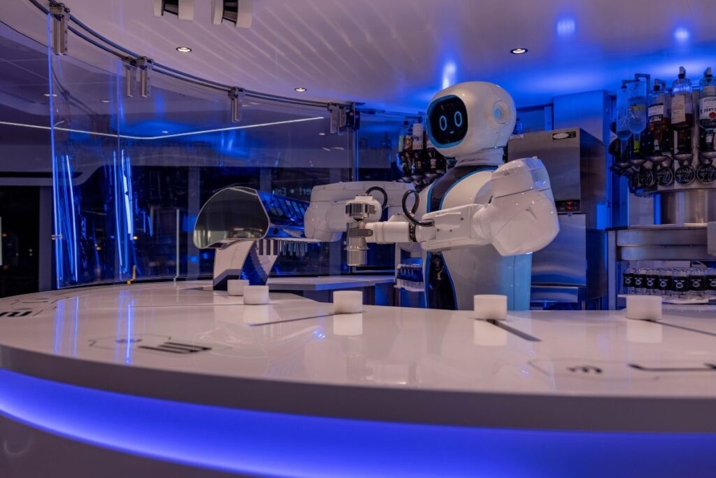 The MSC Starship Club on MSC Virtuosa featuring a robotic bartender in a futuristic setting. The robot, equipped with articulated arms, is positioned behind a sleek, white circular bar illuminated by ambient blue lighting. The area is designed with high-tech elements and transparent, neon-blue accents, creating an innovative and engaging experience for guests.