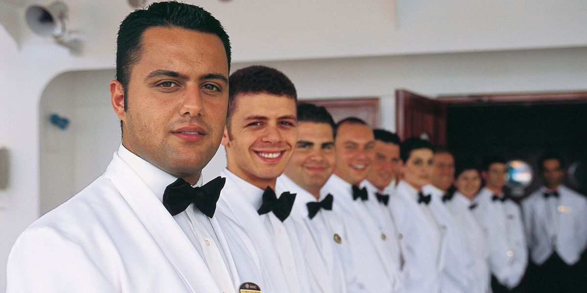 A welcoming group of waitstaff aboard an MSC cruise ship, dressed in crisp white uniforms with bow ties and smiling as they prepare to provide guests with exceptional dining service.
