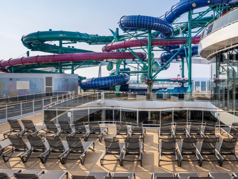 The image shows the deck of a cruise ship featuring multiple large, colorful water slides in shades of blue, green, and purple. In the foreground, there are neatly arranged rows of empty lounge chairs, set up for guests to relax while enjoying the view of the water park area.