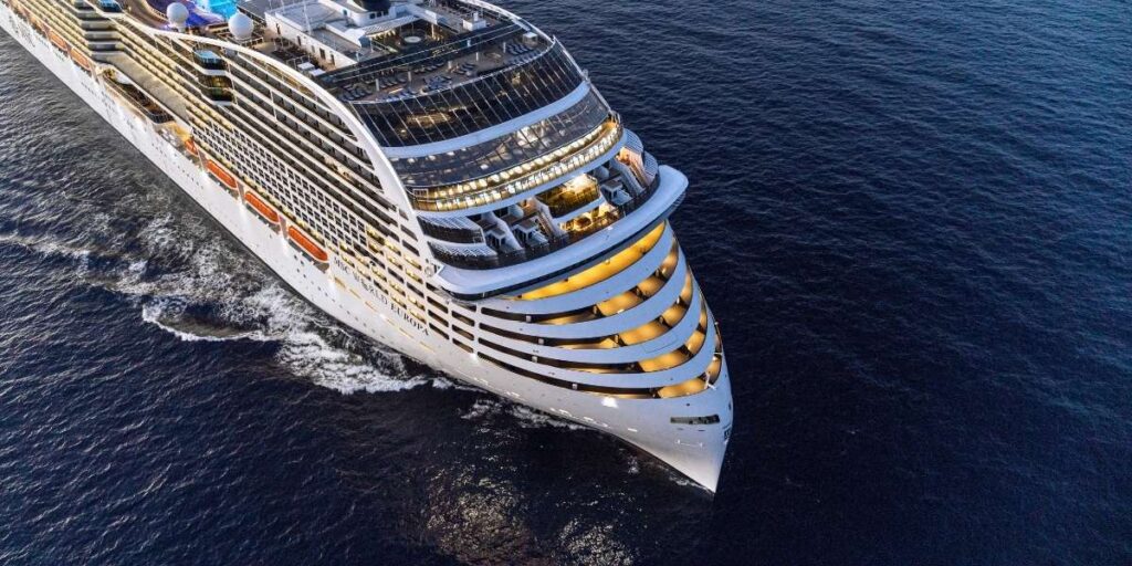 Aerial view of the MSC World Europa cruise ship sailing at sea during twilight, with its curved, futuristic bow design and warmly lit balcony staterooms creating a striking visual. The image showcases the grandeur and innovative architecture of the MSC World Europa cruise ship.