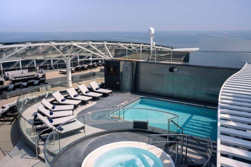 Luxurious sundeck and pool area of the MSC Yacht Club on MSC World Europa. This exclusive space features a modern design with a large infinity pool and a smaller circular jacuzzi, surrounded by white sun loungers. The area is partially covered by a geometric white canopy, offering both sun and shade to guests against a panoramic backdrop of the open sea.