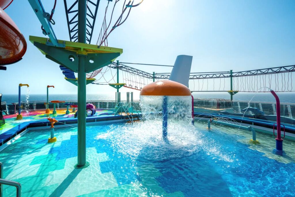 The Aqua Park on MSC Euribia is a splash-filled paradise with a large mushroom water fountain at the center, colorful water play equipment, and a rope bridge against the backdrop of the open sea and blue sky.