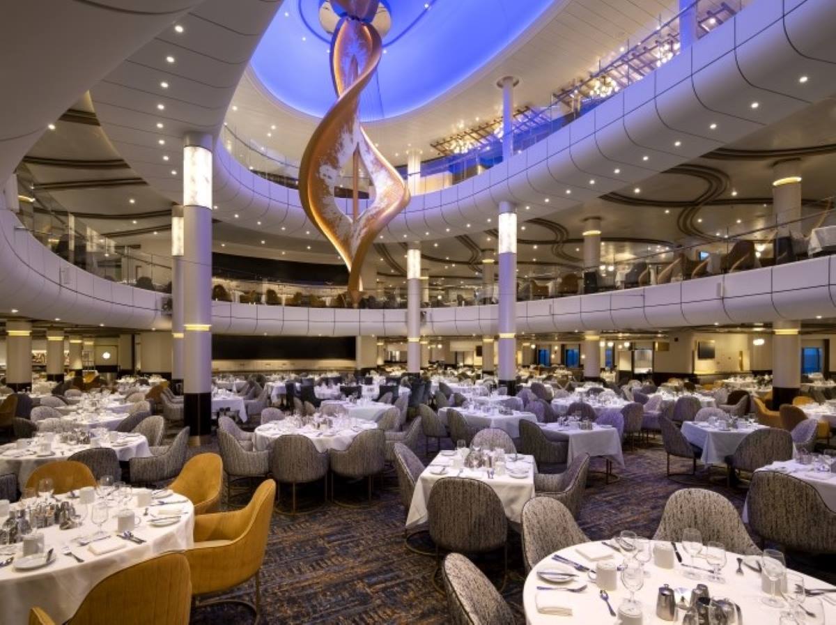The grand Main Dining Room on Spectrum of the Seas cruise ship, displaying a sophisticated decor with circular tiered seating, white linen-covered tables, and a striking copper-toned sculpture under a soft blue light.