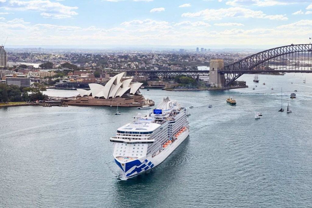 The image features the Majestic Princess cruise ship in the beautiful harbor of Sydney, Australia. The iconic Sydney Opera House with its unique sail-like design and the Sydney Harbour Bridge, known for its arch-based structure, are both prominently visible, framing the vessel. The image captures the essence of Sydney's maritime allure, with the elegant cruise ship contributing to the dynamic and picturesque waterfront of this world-renowned city.