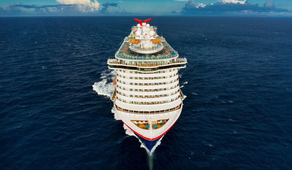 Aerial view of the Mardi Gras cruise ship cutting through the deep blue ocean, showcasing multiple decks and the ship's distinctive red and white funnel.