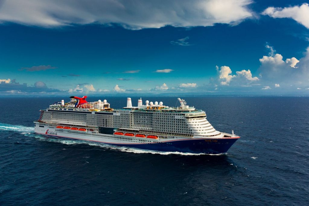 A Carnival cruise ship, the Mardi Gras, sails the deep blue waters under a partly cloudy sky, showcasing its multiple decks and distinctive red funnel.