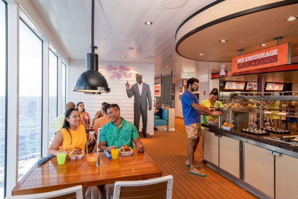 Guests enjoying a meal at Shaq's Big Chicken on Carnival Mardi Gras, a vibrant eatery with ocean views, featuring a casual dining atmosphere with a fun basketball theme and a sign encouraging 'sauciness'.