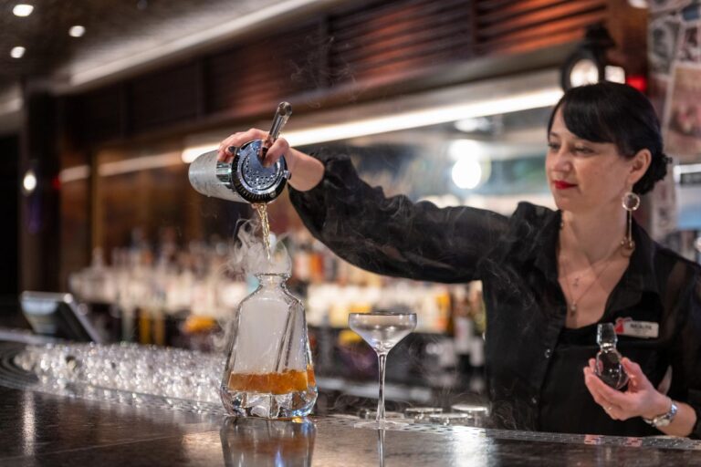 In the image, a bartender at Carnival Mardi Gras' Fortune Teller Bar expertly pours a smoky cocktail into an ornate decanter, showcasing the dramatic flair of mixology aboard the cruise. The bar's atmosphere is enhanced by the bartender's focused expression and the striking visual of the vapor-like effect, promising a mystical and immersive drinking experience.