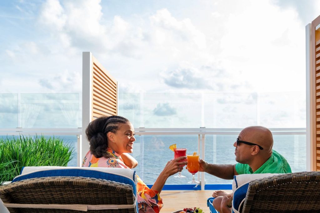 A woman and man in summer attire enjoy tropical drinks on a sunny cruise ship balcony at Loft 19, with the ocean in the background and a clear blue sky above.