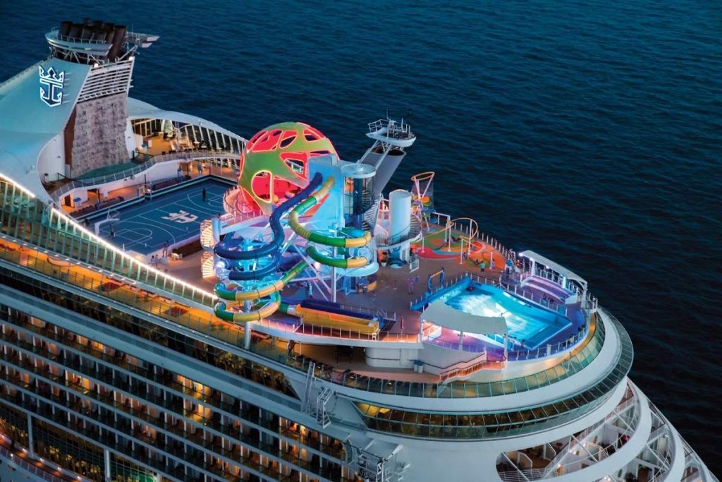 Overhead view at dusk of Royal Caribbean's Mariner of the Seas' sports deck, featuring brightly colored water slides, a rock-climbing wall, and illuminated pools, with the endless ocean extending into the horizon.