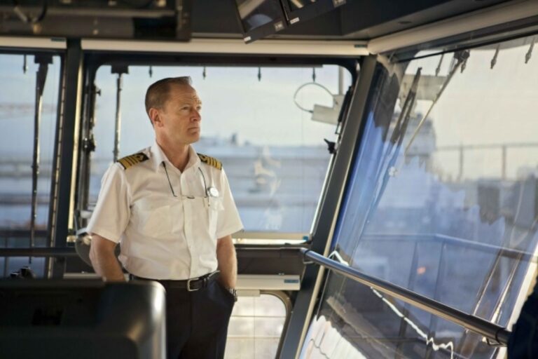 This image features a cruise ship captain standing in the bridge of the ship, dressed in a white uniform with gold-striped epaulettes, gazing out through large windows. The modern interior of the bridge, complete with equipment and panoramic views, emphasizes the captain's role in navigating the vessel. The scene conveys professionalism and the importance of leadership on a cruise ship.