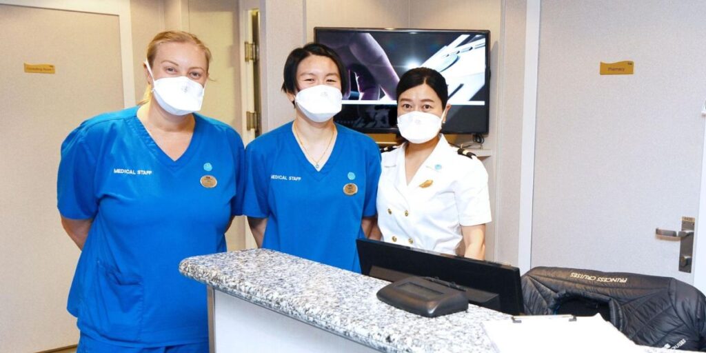 Three cruise ship medical staff members wearing masks and uniforms—two in blue scrubs and one in a white officer’s uniform—stand behind the reception counter of the ship’s medical center. The professional setting reflects the preparedness and care provided by cruise ship medical staff.