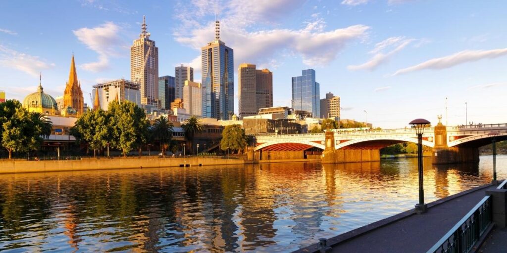 The Melbourne city skyline at sunset with modern skyscrapers, historic buildings, and the Princes Bridge reflected in the calm waters of the Yarra River.