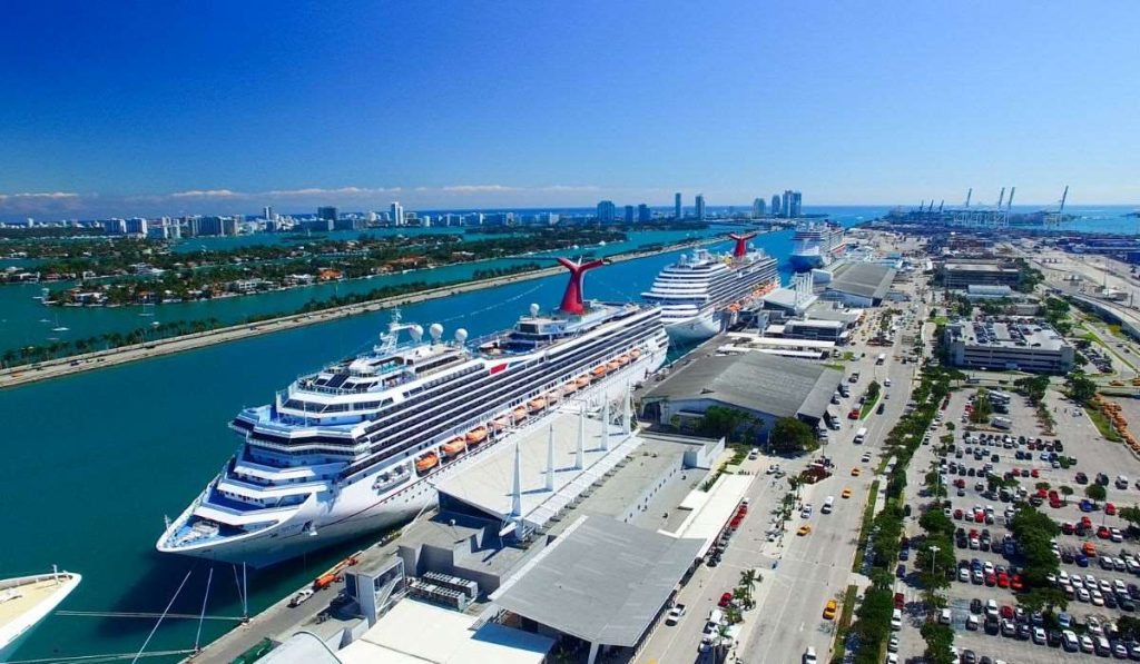 Two Carnival cruise ships docked at the Port of Miami, with iconic red funnels, set against the Miami skyline and clear turquoise waters, under a brilliant blue sky.