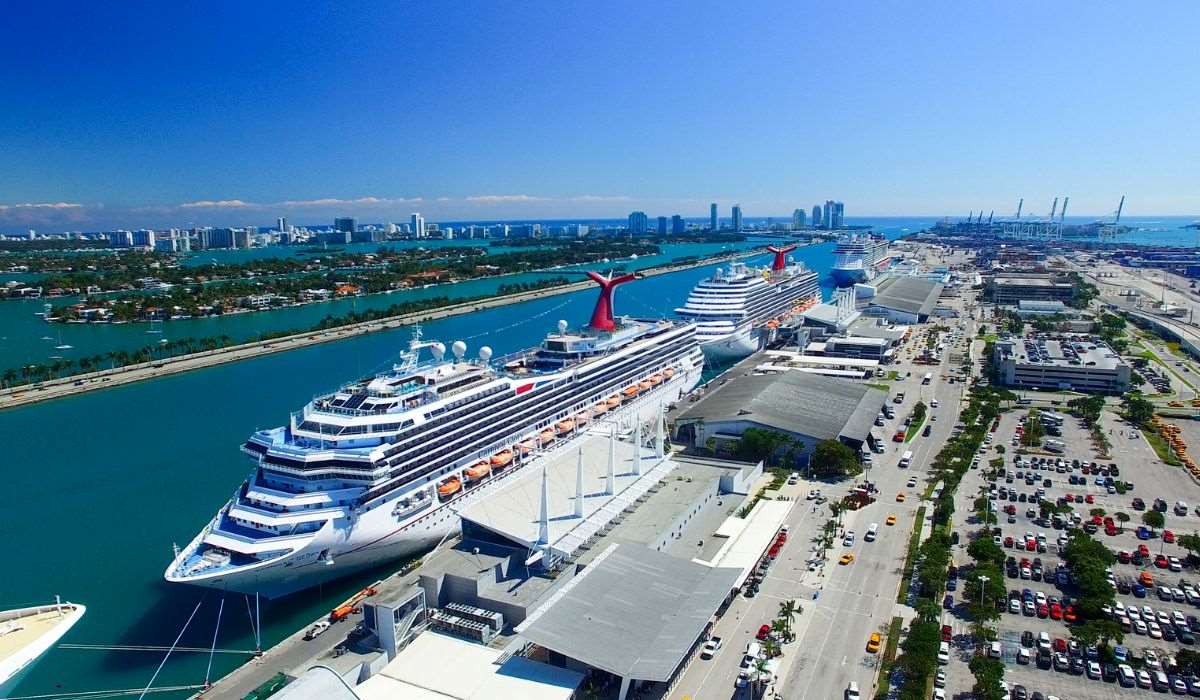 Two Carnival cruise ships docked at the Port of Miami, with iconic red funnels, set against the Miami skyline and clear turquoise waters, under a brilliant blue sky.