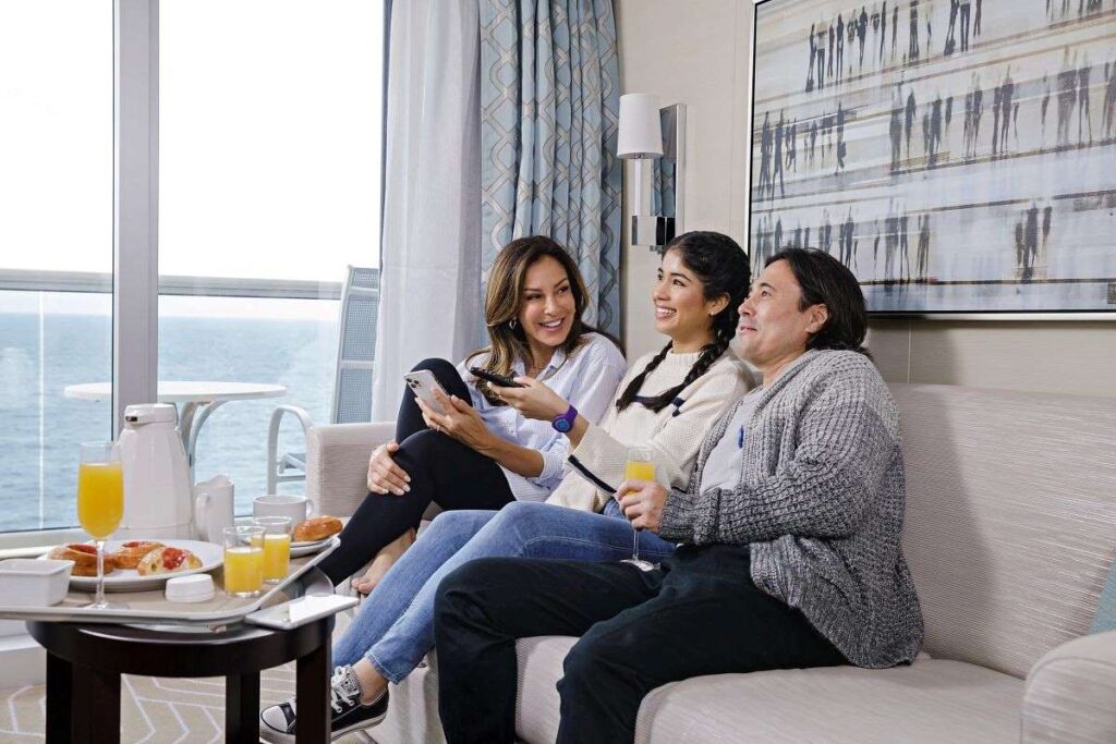 The image depicts a warm and inviting Mini Suite on the Discovery Princess cruise ship. It features a family enjoying a relaxed moment together in a well-appointed room with a contemporary design. Natural light floods in through the large window offering a view of the sea, enhancing the cozy ambiance. The breakfast spread on the table and the comfortable seating arrangement suggest a leisurely morning, with the room’s elegant decor providing a backdrop for a luxurious cruise experience.