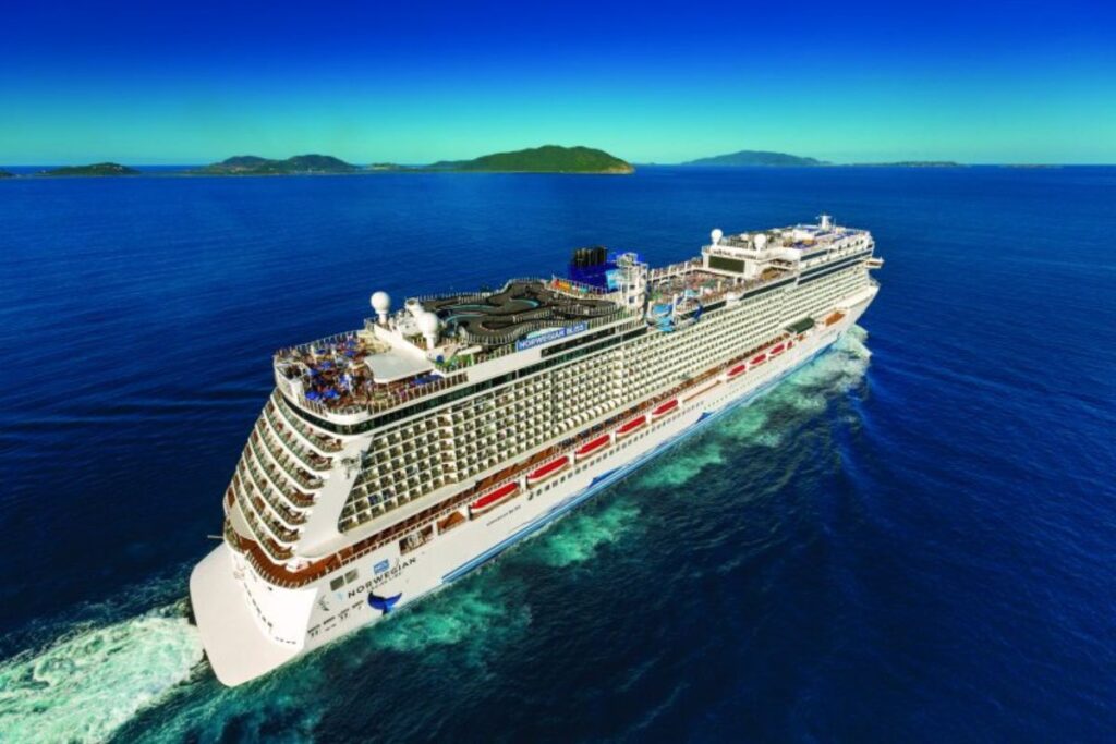 The Norwegian Cruise Line Bliss sails through clear blue waters, with green islands visible in the distance. The ship's expansive decks are filled with amenities, including pools and lounging areas, showcasing its luxurious and spacious design against a stunning seascape backdrop.