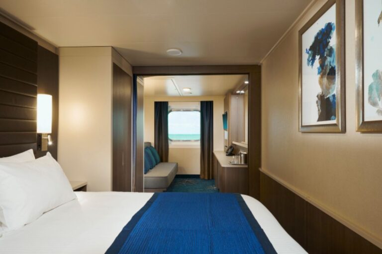 A family ocean view cabin on the NCL Bliss cruise ship features a spacious layout with a large bed, a sofa, and a window offering a view of the ocean. The room is decorated with modern furnishings, including blue accents, a flat-screen TV, and framed artwork on the walls, creating a comfortable and stylish environment for families.