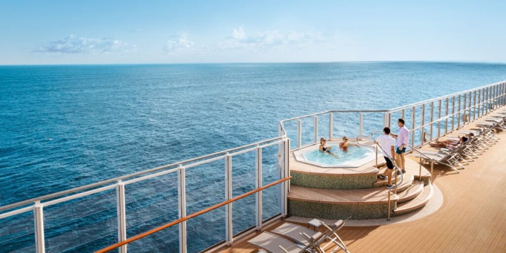 Two people relax in a raised hot tub on the edge of a cruise ship deck, with two more individuals standing nearby enjoying the ocean view. Lounge chairs line the teak deck, and glass panels provide safety while maintaining panoramic views of the open sea.