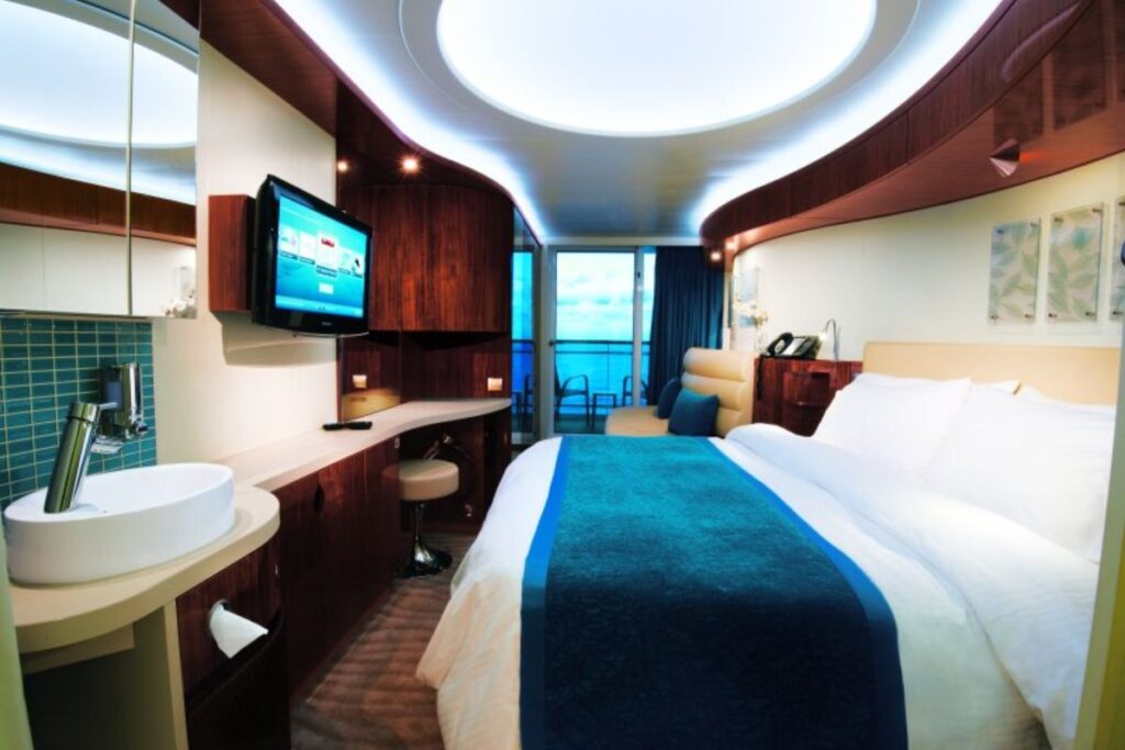 View of a Norwegian Cruise Line Epic stateroom with a balcony, featuring a queen-size bed with white and blue linens, an exposed round basin sink, a wall-mounted TV, and floor-to-ceiling windows leading to the balcony with ocean views.