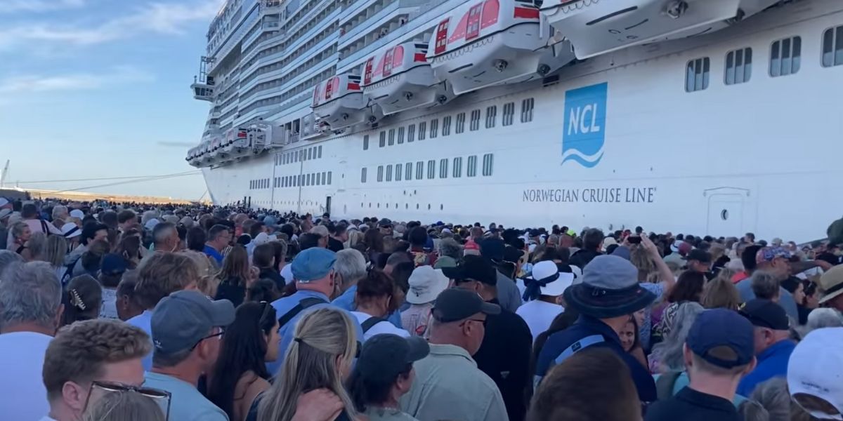 A massive crowd of cruise passengers waits in line beside a Norwegian Cruise Line ship docked at port, creating a packed and congested boarding or disembarkation scene. The NCL logo is visible on the ship’s hull, emphasizing the scale and popularity of Norwegian Cruise Line sailings.