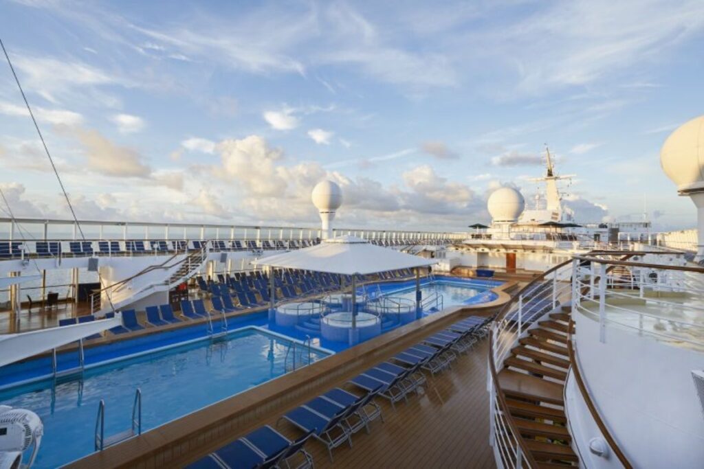 The pool deck on the NCL Sun cruise ship features multiple pools, hot tubs, and rows of blue lounge chairs arranged around the deck. The area is open and spacious, with a clear view of the horizon, providing a perfect setting for relaxation under the sun. Large spherical structures and shaded areas add to the deck's amenities, enhancing the overall experience for guests.