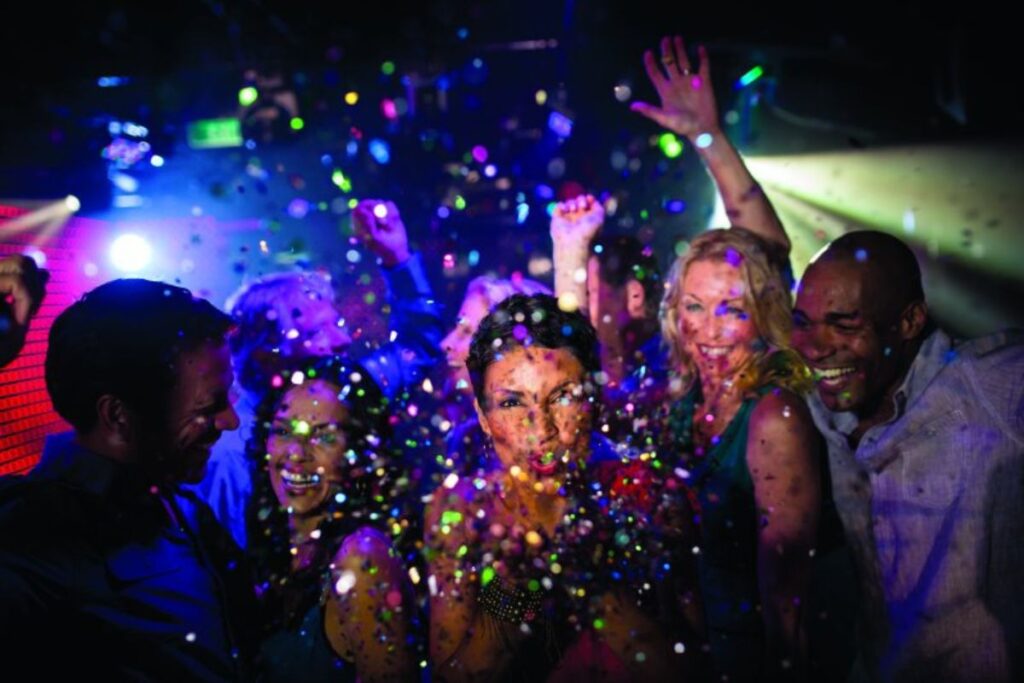 Guests on an NCL cruise enjoy a lively party, with colorful confetti falling around them. The atmosphere is vibrant with flashing lights, and the crowd is smiling and dancing, capturing the energetic and festive spirit of the cruise nightlife.