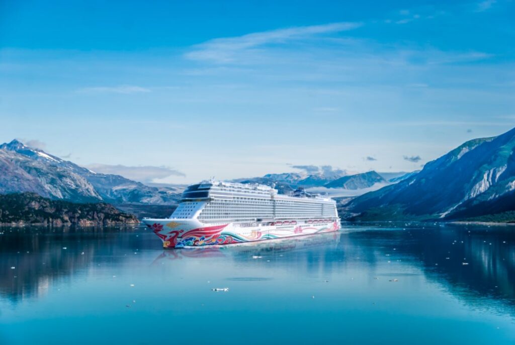This image showcases the Norwegian Joy cruise ship sailing through serene, icy waters surrounded by towering mountains and a clear blue sky. The vibrant hull art contrasts beautifully with the pristine natural environment, emphasizing the ship's modern design amidst the rugged beauty of the landscape. The scene evokes a sense of adventure and luxury in a remote, picturesque setting.