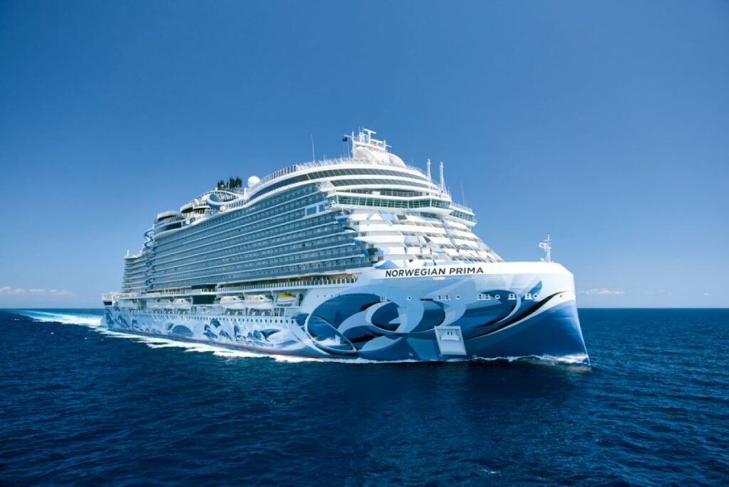 This image showcases the Norwegian Prima cruise ship cutting through the deep blue ocean under a pristine, clear sky. The ship’s hull features sleek, wave-inspired designs in shades of blue and white, emphasizing its modern and elegant aesthetic. Its expansive decks and luxurious build highlight the grandeur and innovation of this state-of-the-art vessel.