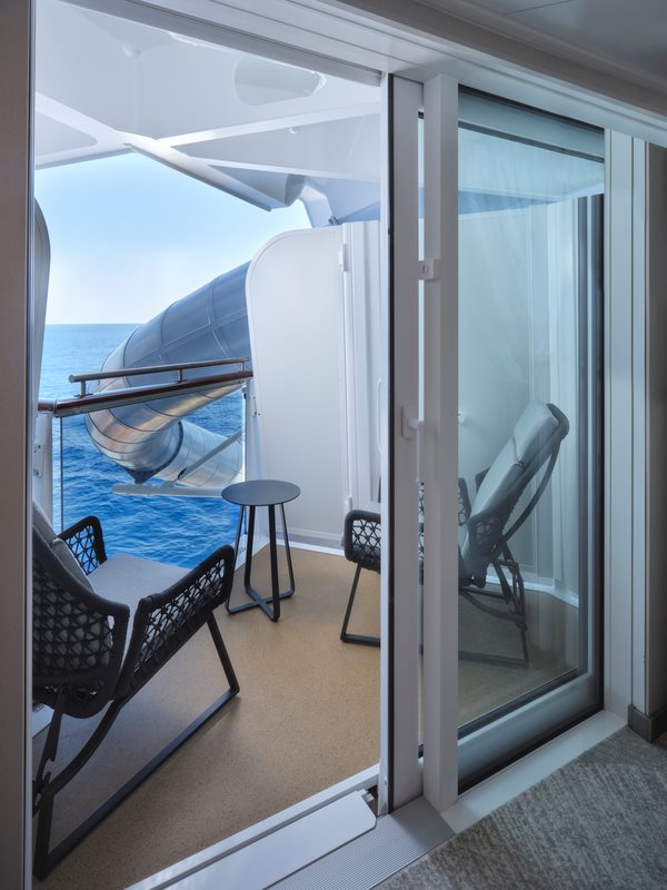 An obstructed oceanview balcony on the Norwegian Prima features a cozy seating area with two chairs and a small table, providing a relaxing spot to enjoy the sea breeze. The balcony has a partial view of the ocean, with a water slide structure partially blocking the view. The space is accessed through sliding glass doors, allowing natural light to fill the cabin.