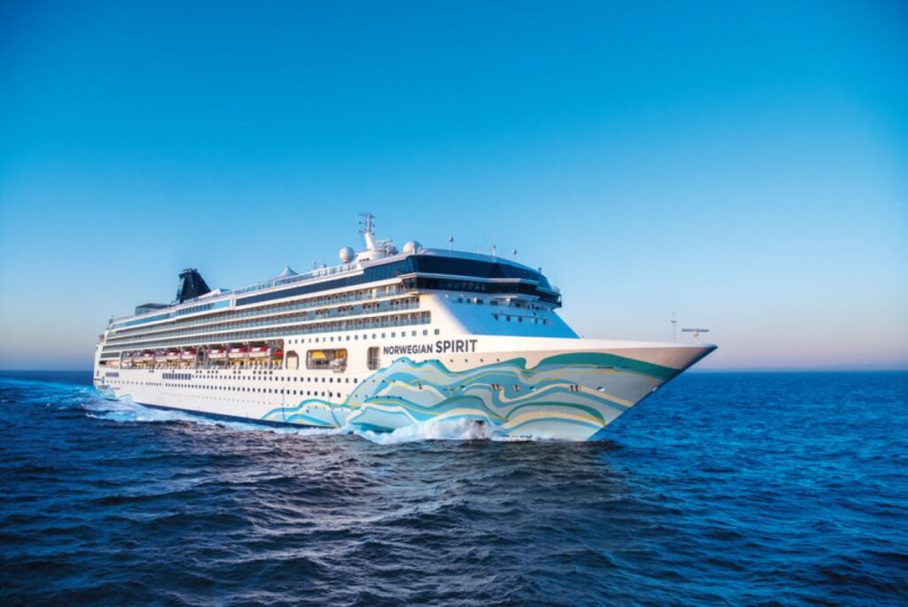 This image shows the Norwegian Spirit cruise ship sailing across calm, deep-blue waters under a clear sky. The hull features elegant wave-themed artwork in shades of blue and green, complementing the maritime setting. The ship's streamlined design and the tranquil sea create a sense of elegance and peaceful travel.