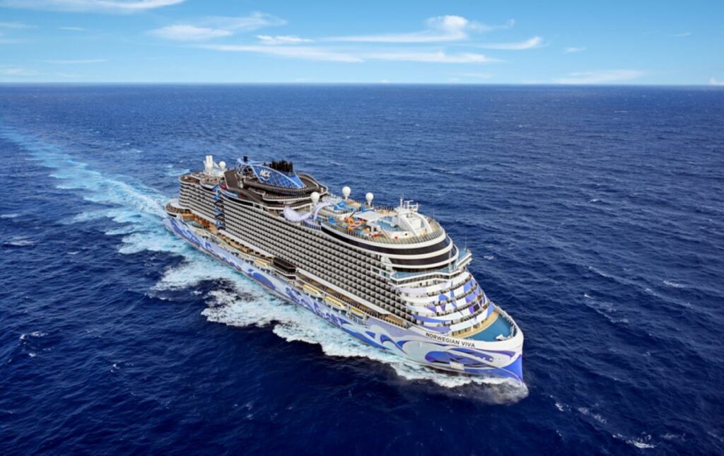 This image features the Norwegian Viva cruise ship gliding through vast blue ocean waters under a clear sky. The ship's hull displays a sleek, wave-inspired design in shades of blue and white, complementing its modern and luxurious aesthetic. The open sea and vibrant sky enhance the sense of freedom and adventure associated with cruising.