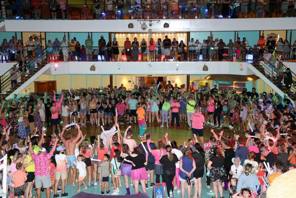 Large crowd of cruise guests gathers on multiple levels around a central dance floor during an 80s-themed Rock-N-Glow party aboard a Carnival Cruise ship. People of all ages, dressed in neon outfits and waving glow sticks, enjoy the festive energy as a Carnival crew member leads the celebration.