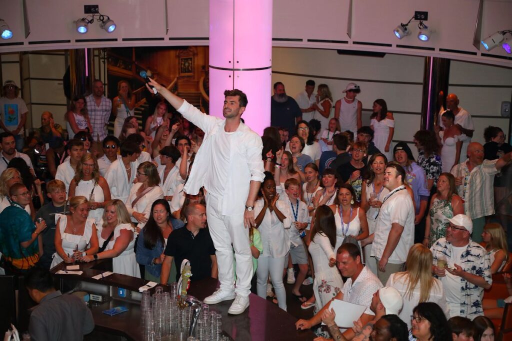 A lively crowd dressed in all-white attire surrounds a man standing on a bar with a microphone, leading a high-energy moment during a White Party aboard a Carnival Cruise. Guests cheer and sing along under colorful lighting in a packed indoor venue.