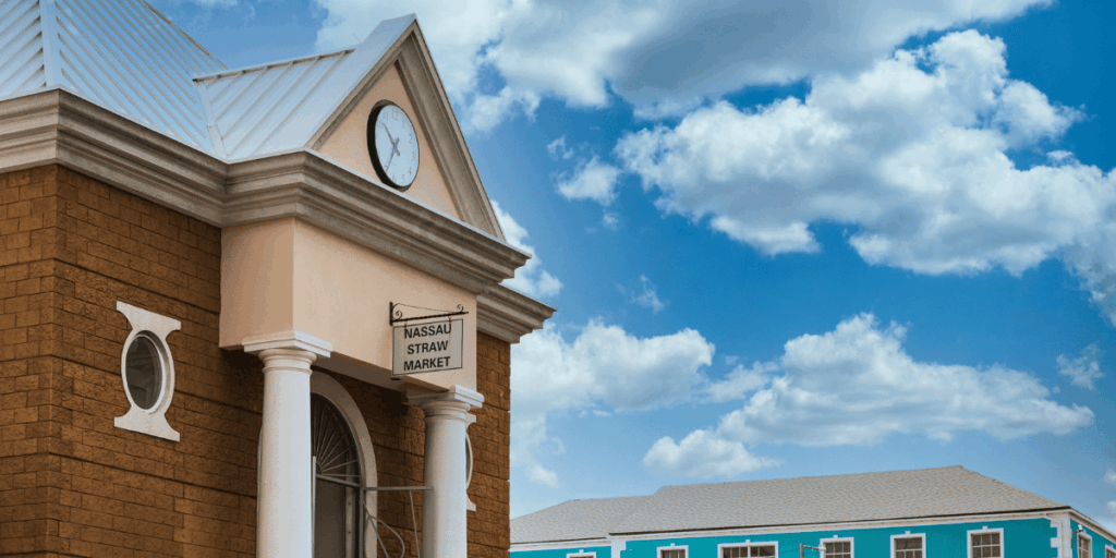 The Nassau Straw Market building features a clock tower with white columns and tan brick walls, set against a bright blue sky with scattered fluffy clouds. A small hanging sign marks the entrance to the popular Bahamian craft market.