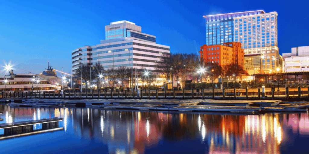 Evening view of downtown Norfolk, Virginia, featuring a mix of modern high-rise buildings and historic brick architecture lit up against a deep blue sky. The waterfront is calm, with empty docks reflecting the city lights along the Elizabeth River.
