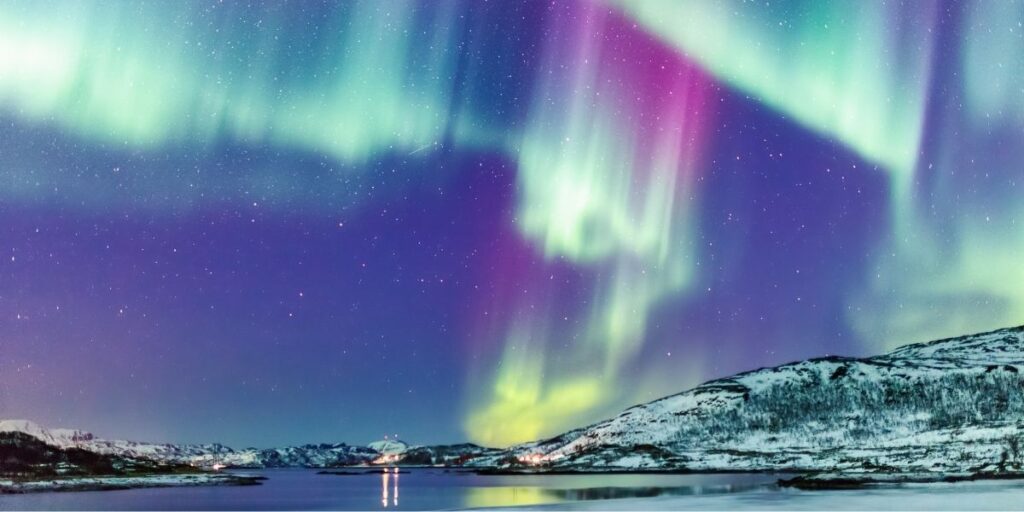 Vivid northern lights in green, purple, and pink hues dance across a star-filled Arctic sky, reflecting over a calm, icy fjord surrounded by snow-covered mountains. The lights from a small settlement glow faintly in the distance beneath the aurora.