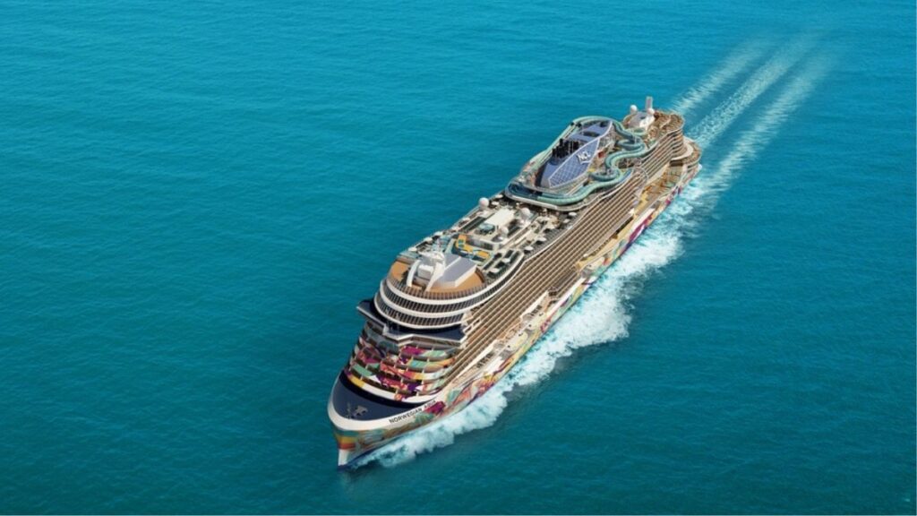 Aerial view of the Norwegian Aqua, a massive and vibrant cruise ship sailing through the open ocean, leaving a gentle wake behind. The ship's colorful hull art stands out against the deep blue sea, while its top deck features water slides, pools, and various attractions. The sleek, modern design highlights the ship’s luxurious amenities and entertainment options.