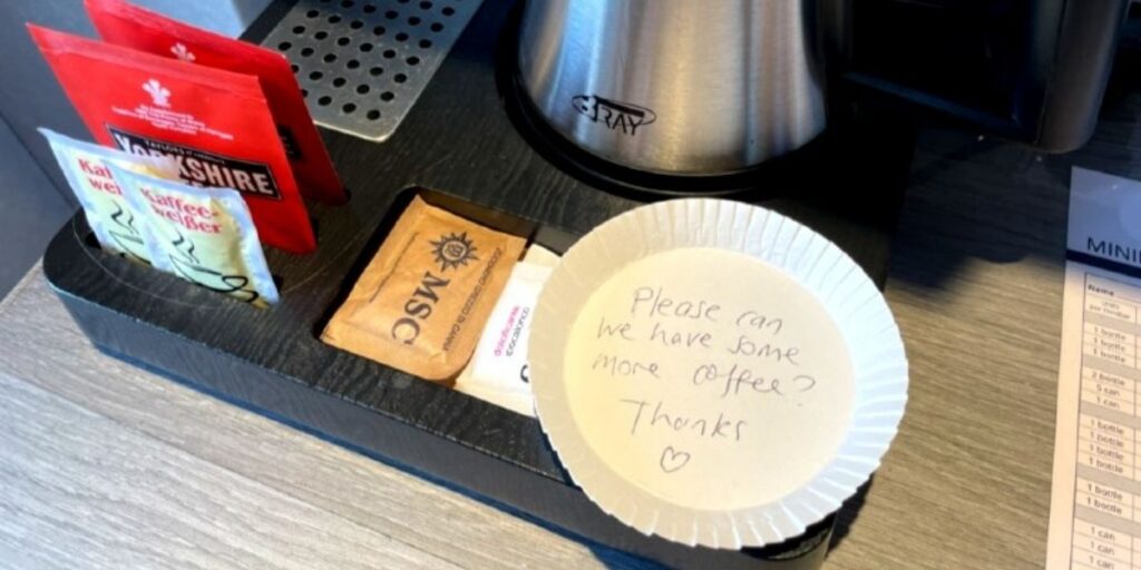 A small cruise cabin coffee station with a few sachets of Yorkshire Tea, coffee creamer, and MSC-branded sugar packets. A paper plate holds a handwritten note reading “Please can we have some more coffee? Thanks ♥,” left neatly beside a silver kettle.