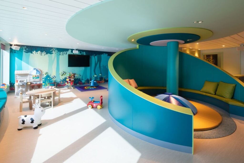 The image displays a vibrant and child-friendly nursery on a P&O cruise ship. The room is brightly lit, featuring colorful decor with ocean-themed wall murals, a small seating area, and various toys like a rocking cow and play kitchen. The design emphasizes safety and fun, with soft rounded structures and engaging activities for young children.