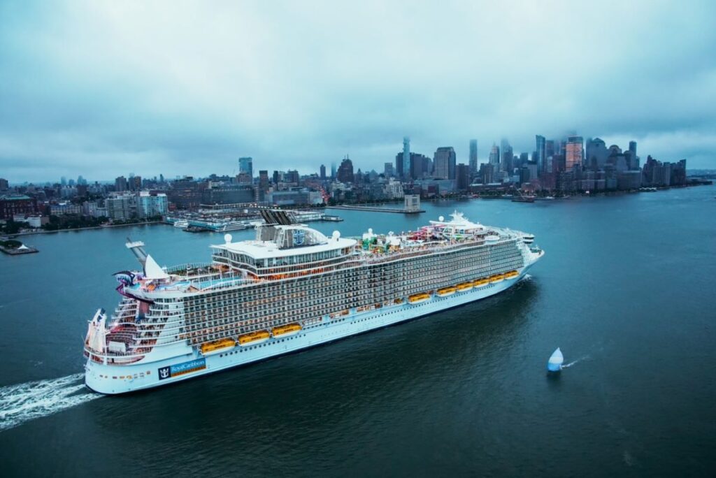 Overlooking a dreary New York City skyline, Royal Caribbean's Oasis of the Seas presents a contrasting image of leisure and adventure on the Hudson River, its vibrant design elements standing out against the muted urban backdrop on a cloudy day.