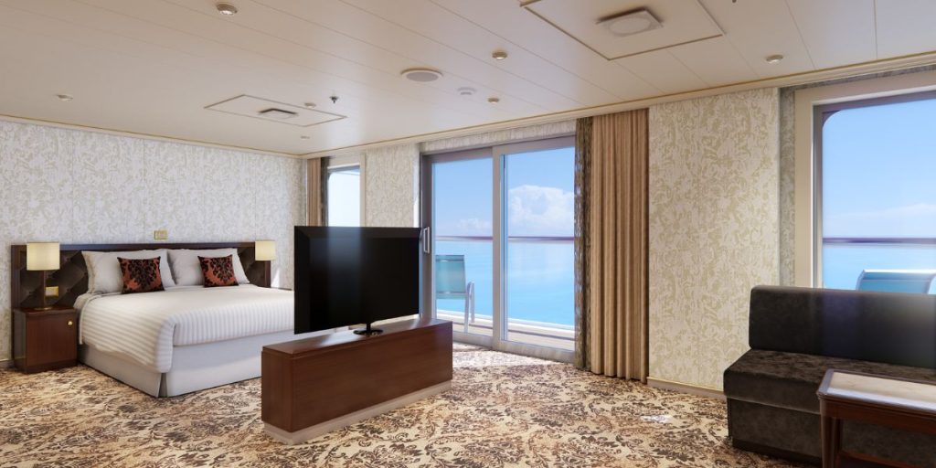 An Ocean Suite with a classic design, featuring a plush bed with decorative pillows, an ornate wallpapered accent wall, and a luxurious patterned carpet. The suite includes modern amenities like a large flat-screen TV and offers a spectacular ocean view through the sliding glass door that leads to a private balcony. The room's elegant furnishings and the tranquil sea view provide a perfect blend of comfort and sophistication for a relaxing cruise experience.