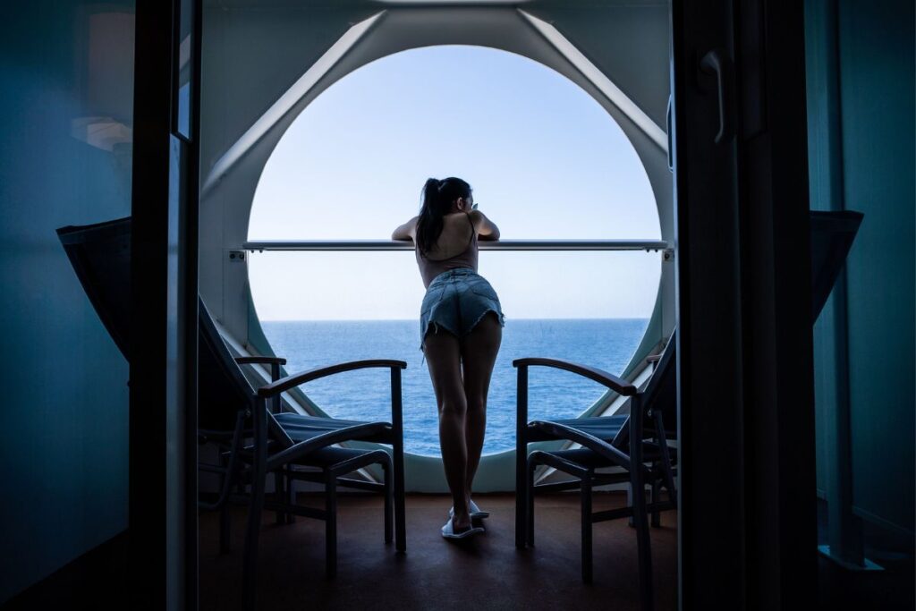 A person stands on the balcony of a cruise ship, gazing out at the ocean. The silhouette framed by the arched balcony doorway contrasts with the tranquil blue sea, capturing a moment of peaceful contemplation on the open waters.