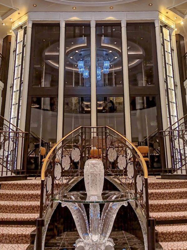 The image shows a grand staircase aboard the Oceania Mariner, featuring ornate railings with intricate patterns. At the base of the stairs is an elegant glass sculpture, serving as a focal point. The backdrop reveals tall, floor-to-ceiling windows and a beautiful chandelier, adding a luxurious and sophisticated ambiance to the space.