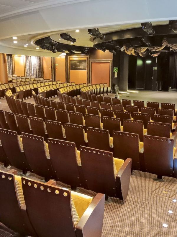 This image shows the theater on the Oceania Mariner cruise ship. It features rows of neatly aligned wooden chairs with cushioned seating, arranged in a semicircle around a small stage. The stage is framed by elegant drapes, and the lighting system is visible overhead. The intimate design of the theater suggests it is intended for live performances, talks, or presentations during the cruise.