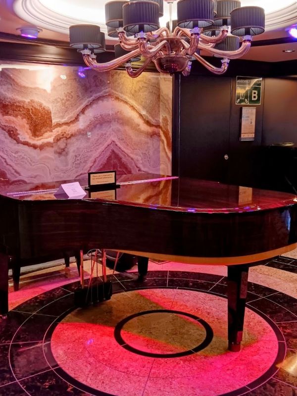 The image shows a grand piano on the Oceania Mariner, placed on a glossy, circular marble floor. Above the piano is an elegant chandelier with multiple black lampshades, casting soft lighting. The backdrop features a stunning marble wall with natural, swirling patterns in warm tones, contributing to the luxurious ambiance of the space, likely intended for live performances or a lounge setting.