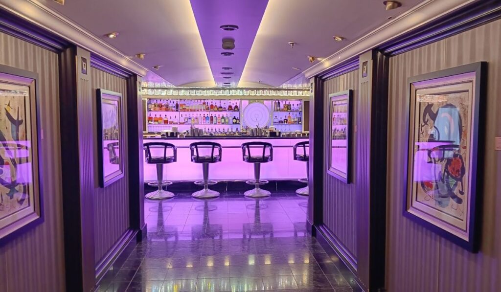The image shows a sleek bar area on the Oceania Mariner, with modern bar stools and a backlit bar displaying a wide array of liquor bottles. The lighting is a soft purple, creating a contemporary, elegant ambiance. The hallway leading to the bar is adorned with framed art, adding a sophisticated touch to the setting.