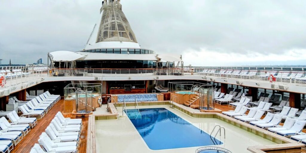 The image shows the pool deck of an Oceania cruise ship. The scene features a rectangular swimming pool in the center, surrounded by wooden decking and rows of cushioned lounge chairs neatly lined up. Two jacuzzi areas are visible near the pool, and the large funnel structure dominates the background. The ship's deck is designed for relaxation, with ample seating and an open, spacious layout under cloudy skies.